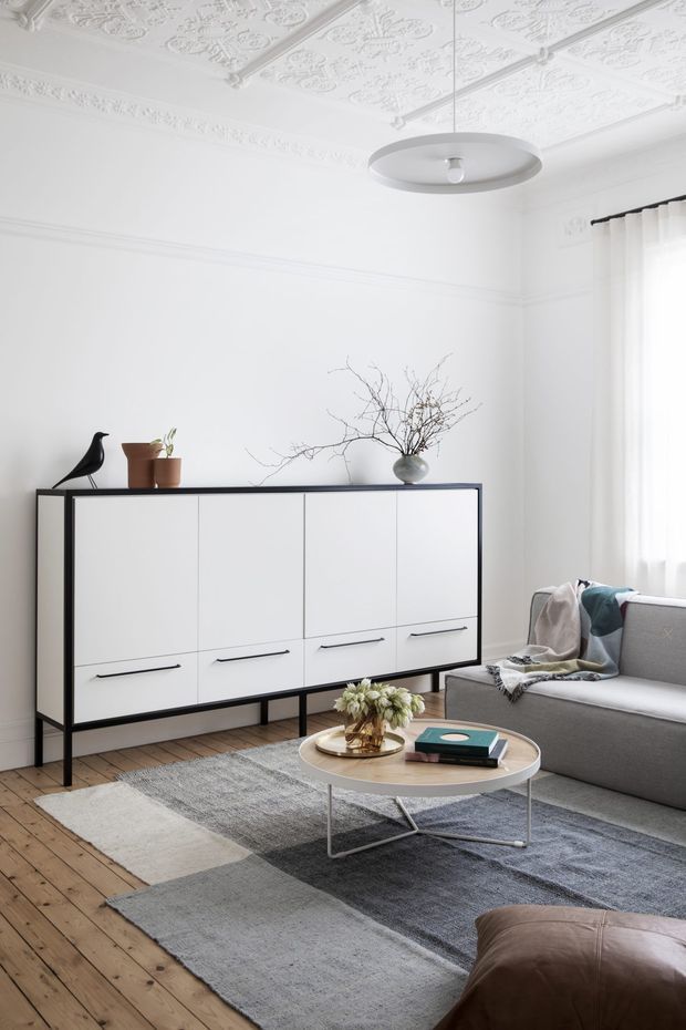 Bright, refined living room in a period home, featuring an ornate white pressed-tin ceiling and decorative cornice, white walls, timber floorboards, and sheer white curtains. A contemporary white and black sideboard sits against the wall beside a grey upholstered sofa. Layered grey rugs, a round light coffee table, simple pendant light, and restrained botanical and sculptural decor create a calm modern-classic interior.