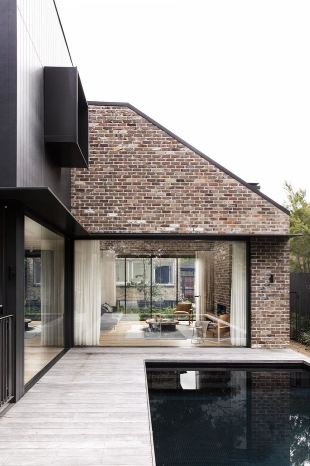 Exterior view of a contemporary courtyard-style residence combining dark metal cladding and framing with a prominent recycled-looking brick gable wall. Full-height glazing opens the living area onto a pale timber deck, with a rectangular reflecting or swimming pool in the foreground. The design has a strong geometric composition, clean lines, and a high-contrast black, brick, and light timber palette.