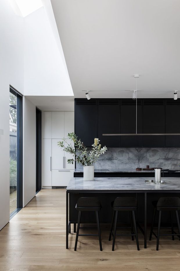 A contemporary kitchen with a large black island and integrated dark cabinetry, three black bar stools, and a dramatic grey-veined stone benchtop and splashback. White cabinetry and pale timber flooring balance the dark joinery. The room is bright and open, with a high ceiling, clerestory-like glazing, and a slim linear pendant above the island.