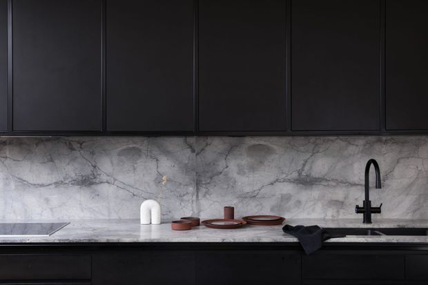 Close-up view of a sophisticated dark kitchen with full-height matte black cabinetry, a dramatic grey veined stone splashback and benchtop, and a black gooseneck tap over an undermount sink. Minimal ceramic and dark-toned servingware sit on the benchtop, creating a refined, moody composition.