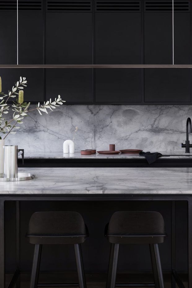 A dramatic, dark-toned contemporary kitchen with matte black full-height cabinetry, integrated horizontal ventilation detailing, a veined grey marble splashback and island benchtop, and a black undermount sink with a tall black tap. Two dark bar stools sit at the island, while restrained ceramic tableware and a simple branch arrangement add subtle styling.