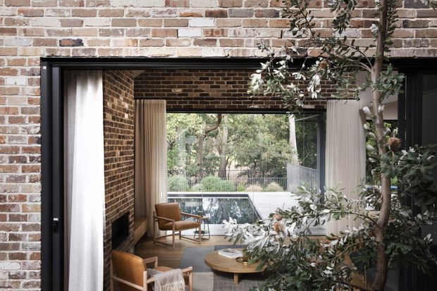 View through a large black-framed opening in a warm exposed-brick interior toward a glazed outdoor pool area. The room contains timber flooring, mid-century style wooden armchairs, a round wooden coffee table, curtains, and a dark brick fireplace wall. Mature planting partially frames the view, creating a strong indoor-outdoor connection.