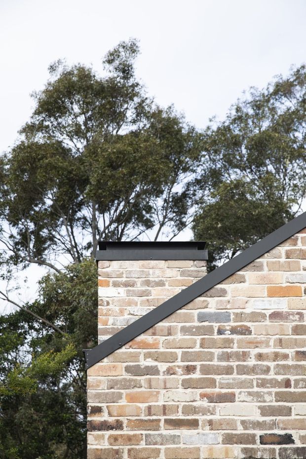 Close-up exterior architectural detail showing a pale multi-toned brick wall with dark metal roof edging and a sloping gable line. Mature trees form the background, emphasizing the building envelope and roof junction.