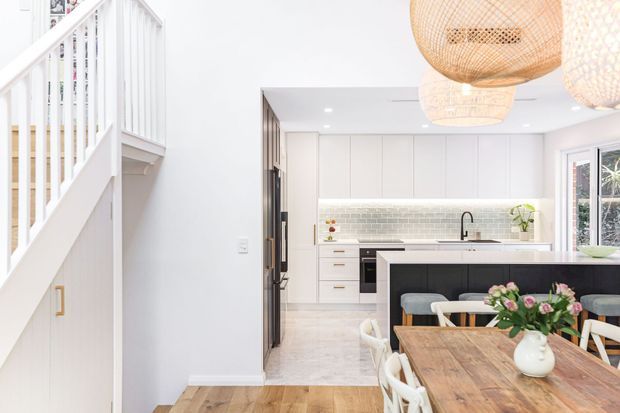 Bright open-plan kitchen and dining area viewed from beside a white staircase. The kitchen features full-height white cabinetry, a pale textured tile splashback, dark island cabinetry with a light benchtop, and a black tap. A timber dining table with white chairs occupies the foreground, while woven pendant lights add a natural decorative element.