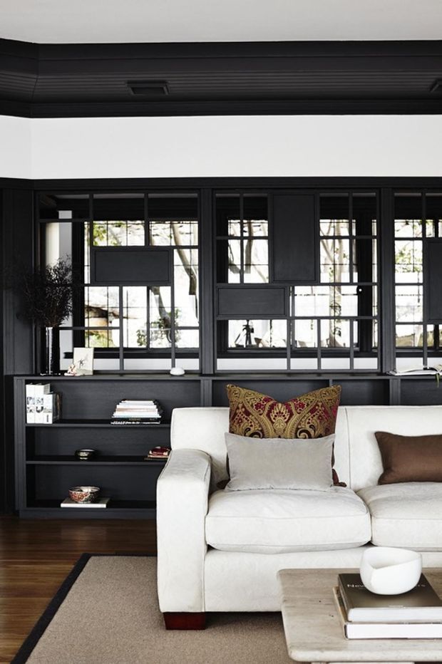 A refined living room featuring a white upholstered sectional sofa, dark timber built-in shelving and cabinetry, and a dramatic black-framed interior window wall with multiple glazed panes and inset opaque panels. A dark coffered or paneled ceiling, warm timber flooring, neutral rug, patterned cushions, and a pale stone or concrete coffee table create a high-contrast, tailored interior.