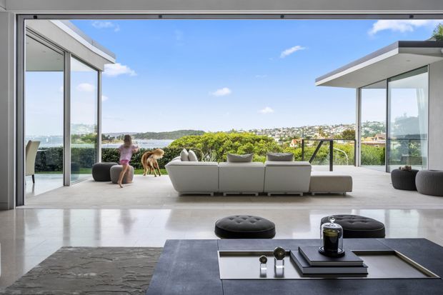 A bright, contemporary open-plan living room extending onto a covered terrace through large floor-to-ceiling sliding glass doors. The room features a pale polished hard floor, a large dark coffee table with books and decorative objects, low upholstered seating, and a broad view toward a green waterfront hillside. Flat roof planes, minimal framing, and strong indoor-outdoor connection define the architecture.