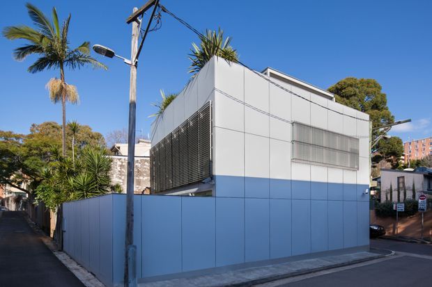 Street-facing view of a compact contemporary flat-roof building with a crisp white upper facade and pale blue lower cladding. The facade features large rectangular panels, horizontal external louvres, a screened window, and rooftop planting or palms. Utility poles, paved streets, neighbouring buildings, and mature vegetation surround the structure in a sunny urban setting.