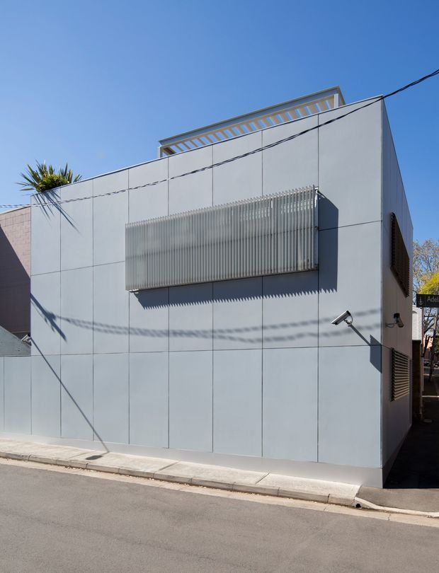 Street-level exterior view of a compact contemporary building clad in smooth pale grey panelled surfaces. The facade features a projecting translucent ribbed screen, louvred side openings, rooftop balustrading, external security lighting, and clean rectilinear geometry on an urban street corner.