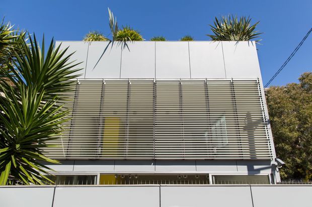 Exterior view of a contemporary two-storey building with a clean white rectangular facade. The upper level is screened by extensive horizontal louvres, with narrow glazing visible behind, while the flat roof includes spiky architectural planting. A solid white boundary wall occupies the foreground, with mature vegetation at the sides and overhead utility wires against a clear blue sky.