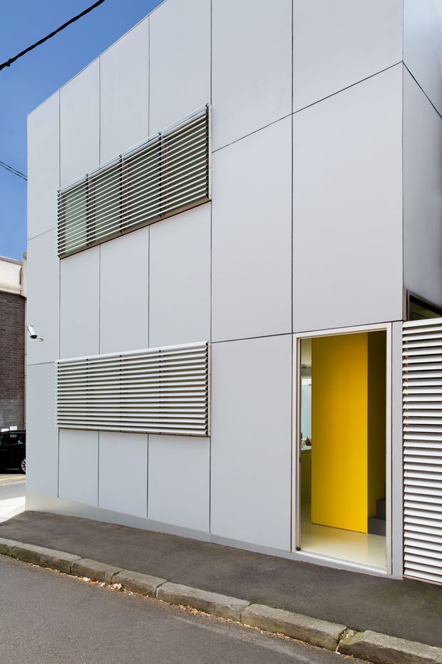 Street-level exterior view of a compact contemporary building clad in large pale grey panels. Horizontal external louvres shade the windows, and an open doorway reveals a vivid yellow interior wall and light floor. The building sits directly beside a narrow paved footpath and road, with neighbouring urban structures partially visible.