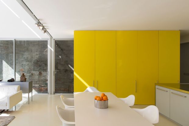 A bright, contemporary open-plan kitchen and dining area with a long white island or dining counter surrounded by white sculptural chairs. A vivid glossy yellow floor-to-ceiling bank of built-in cabinetry forms the main feature wall, while pale cabinetry continues at the right. Full-height glazing and a glass partition reveal a rough grey concrete or stone wall beyond. Track lighting, white surfaces, and strong sunlight create a clean, high-contrast interior.