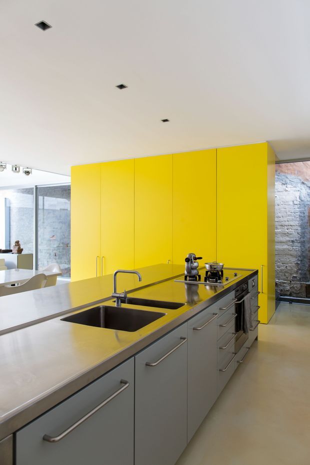 A bright contemporary kitchen dominated by a long central island with an integrated double-basin sink, chrome tapware, grey cabinetry, and a stainless-steel benchtop. Tall vivid yellow full-height cabinetry forms a strong feature wall, while exposed pale brick walls and extensive glazing add an industrial character. A dining area is partially visible beyond the kitchen.