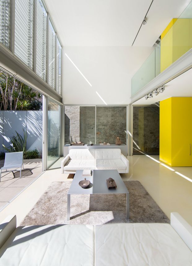 Bright, double-height contemporary living room with expansive louvred glazing, polished pale flooring, white leather sofas, a minimalist grey coffee table, and a neutral rug. A vivid yellow built-in wall and cabinetry provide a strong accent, while a glazed upper-level mezzanine or landing overlooks the room. Exposed textured masonry forms the rear wall, and an adjacent outdoor courtyard is visible through the glazing.