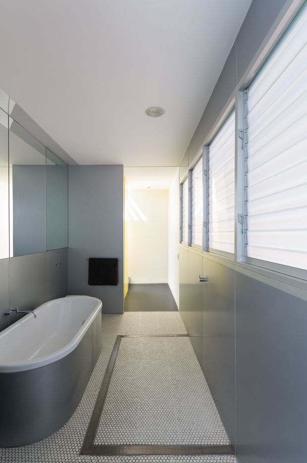 A bright contemporary bathroom with a freestanding white bathtub, grey wall surfaces, full-height mirrored panels, and long horizontal louvred windows. The floor is finished in small light mosaic tiles with darker bordered inset mats, creating a clean, minimalist wet-area design.
