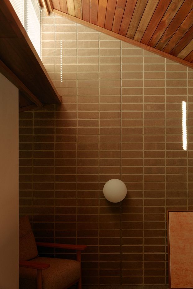 A warm, moody interior detail showing a tall wall of horizontally laid brown brick, a wood-lined sloping ceiling with exposed timber framing, and a spherical wall light. Part of a timber-framed upholstered chair and a framed artwork are visible at the edges. Strong sunlight creates narrow vertical highlights across the wall.
