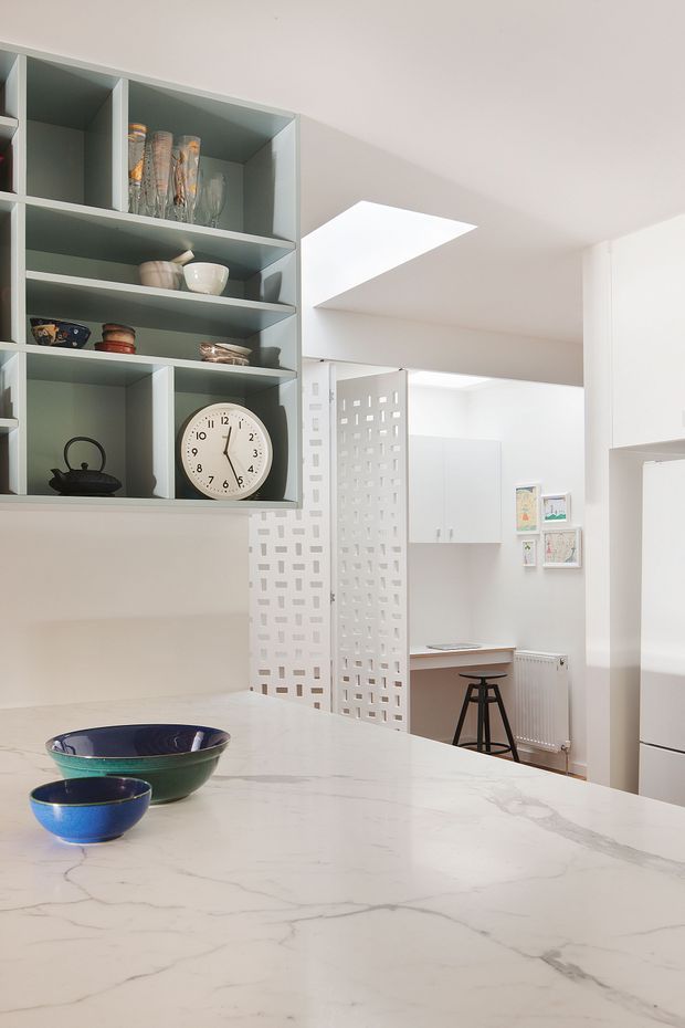 Bright contemporary kitchen interior viewed across a large white veined stone island. Pale blue-grey open shelving displays glassware and ceramics, while a decorative white perforated room divider separates the kitchen from a compact work area. White cabinetry, a skylight, a stool, radiator and framed artwork are visible in the background.