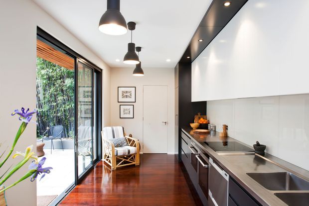 Bright contemporary galley kitchen with dark timber flooring, white walls and cabinetry, black pendant lights, and a long dark benchtop with integrated cooking and sink areas. Floor-to-ceiling sliding glass doors open to a small outdoor terrace, while a rattan lounge chair and framed artwork add warmth. The space has a clean, compact layout with strong indoor-outdoor connection.