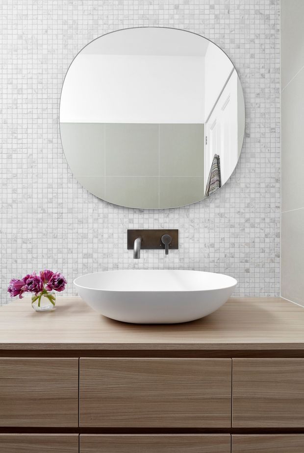 A contemporary bathroom vanity featuring a large round mirror, white vessel basin, wall-mounted chrome tapware, pale mosaic wall tiles, and a timber-look built-in vanity cabinet with a light wood benchtop. A small vase of flowers sits beside the basin.