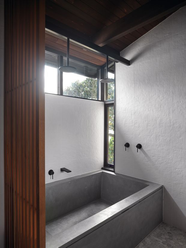 A moody, minimalist bathroom featuring a deep built-in concrete bathtub, dark wall-mounted tapware, textured white plaster walls, black-framed clerestory and vertical windows, and a warm timber slatted screen. The bathroom has a high exposed timber-lined ceiling with dark structural beams and two overhead shower fixtures, combining raw concrete, timber, and natural light.