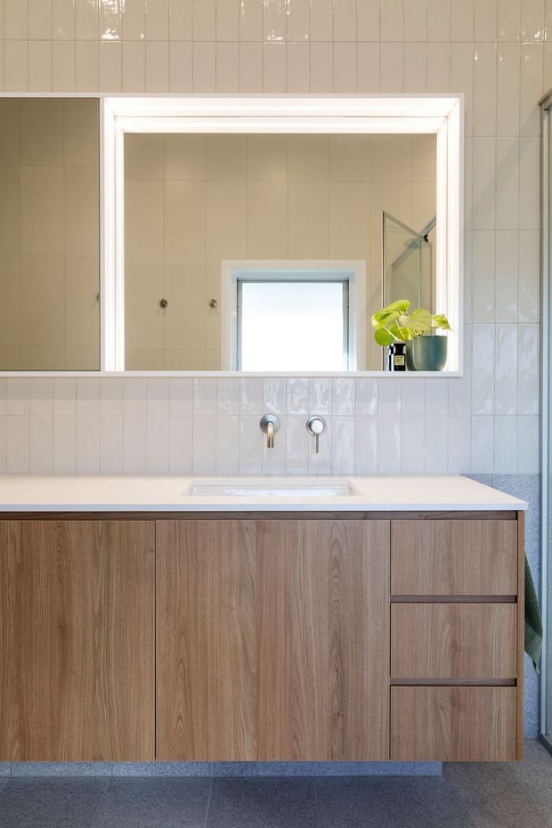 A bright, contemporary bathroom vanity featuring a wall-mounted timber-look cabinet with integrated drawers, a pale stone or engineered-stone benchtop, and a rectangular inset basin. Glossy light ceramic wall tiles surround a large mirrored cabinet, with chrome wall-mounted tapware and a small potted plant adding detail. A shower enclosure is partially visible at the right edge.