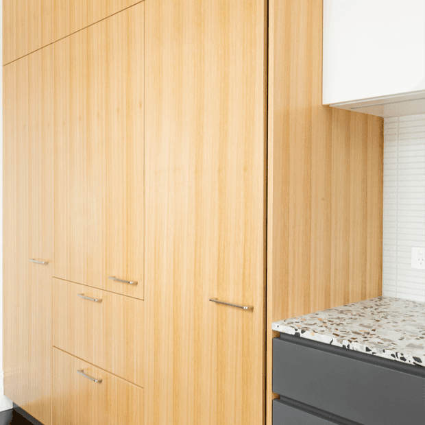 Close-up of a contemporary kitchen or utility space featuring tall light timber veneer cabinetry with slim horizontal metal handles, dark grey lower cabinetry, a terrazzo-style benchtop, and a white tiled splashback. The image focuses on fixed joinery and material detailing.