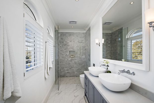 Bright, refined bathroom with twin vessel basins on a grey vanity, a large framed mirror, walk-in glass shower, toilet, marble-look floor tiles, and grey stone-look shower wall tiles. White plantation shutters, arched window detailing, wall sconces, and decorative ceiling mouldings create a classic polished interior.