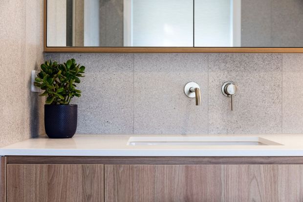 Close-up contemporary bathroom vanity with a wide pale engineered-stone benchtop and integrated rectangular basin, wall-mounted chrome tapware, warm wood-look cabinetry, and a large framed mirror. The grey speckled wall finish and restrained detailing create a clean, refined aesthetic.