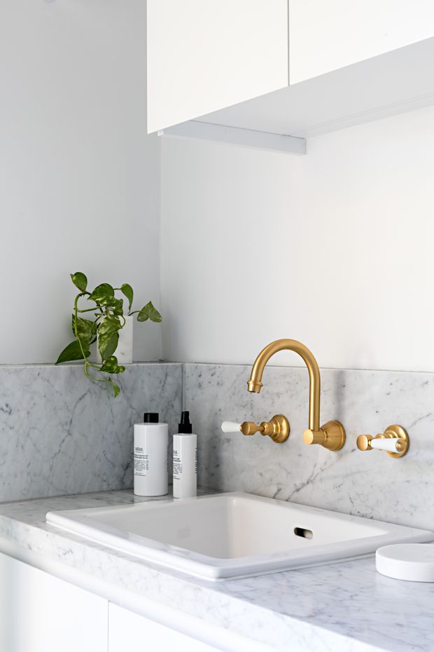 Bright, refined bathroom vanity featuring a white inset basin set into a white-veined marble countertop with matching marble splashback. A polished brass wall-mounted tap set provides a warm accent, while white cabinetry and a small trailing plant complete the minimalist composition.
