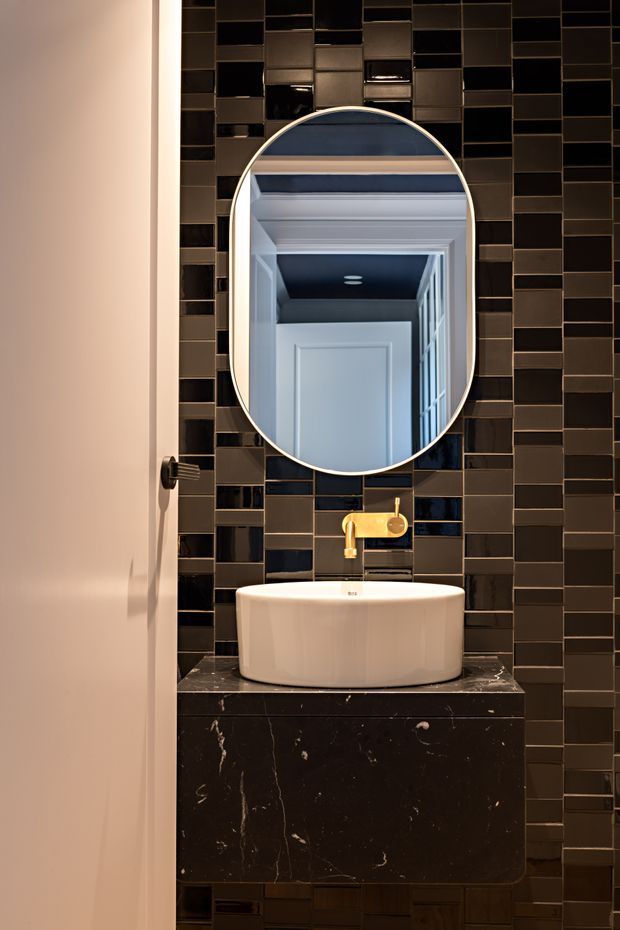 A compact, dramatic bathroom vanity featuring a white vessel basin on a dark marble-look floating vanity. The wall is clad in glossy rectangular dark tiles with contrasting grout, and an arched oval mirror with a slim light frame is mounted above a brass wall-mounted tap. A white paneled door is visible at the left, while the mirror reflects a hallway or adjacent interior with decorative ceiling trim.