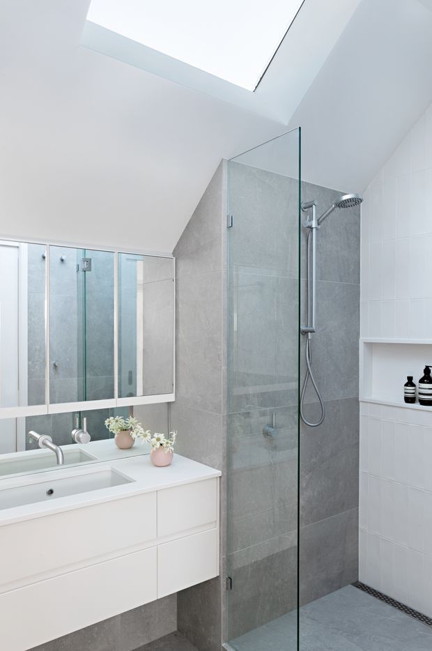 Bright contemporary bathroom with a frameless glass walk-in shower, grey large-format tiled walls and floor, white wall-mounted vanity with integrated basin, mirrored cabinet, recessed shelving, chrome fixtures, and a large skylight. The sloped ceiling and skylight bring abundant natural light into the compact space.