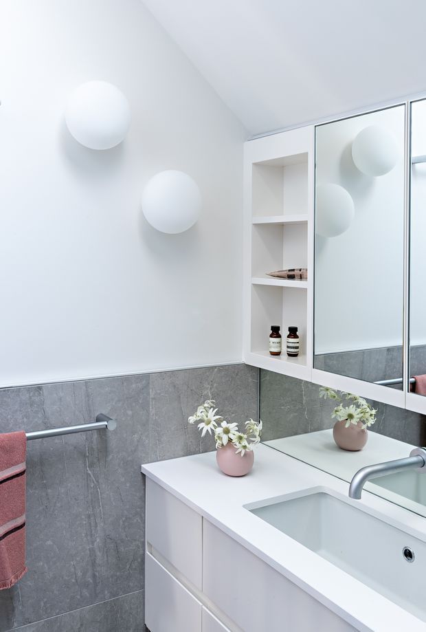 Bright, compact contemporary bathroom vanity area with a white integrated sink and benchtop, pale cabinetry, grey tiled lower walls, a large mirrored wall cabinet with open shelving, spherical wall lights, and a small vase of flowers. A towel rail with a pink towel is visible at left.