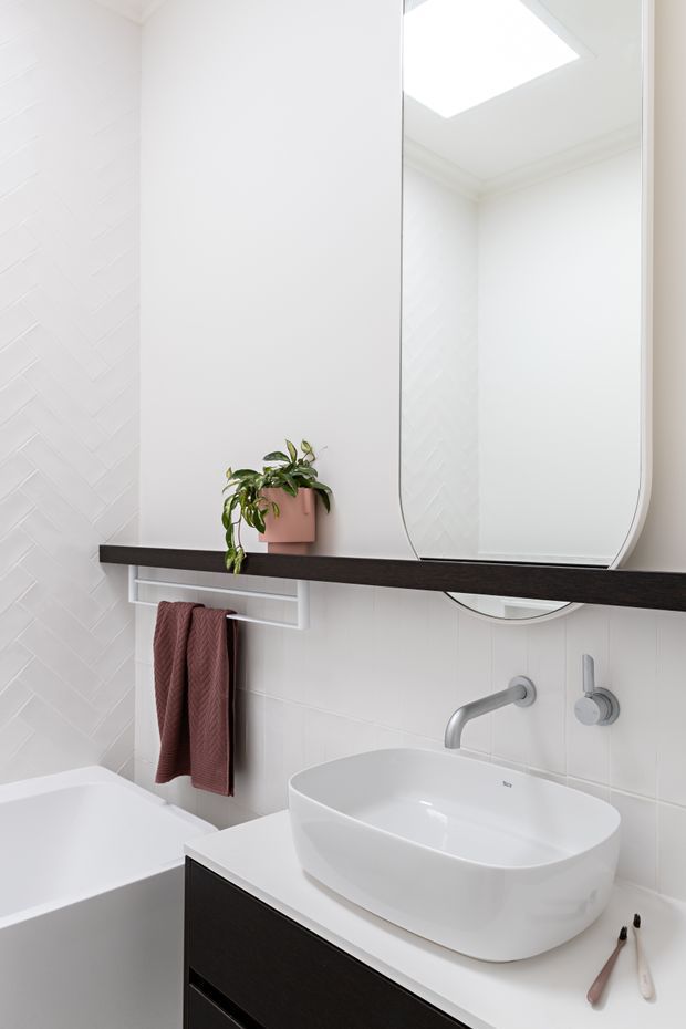 Bright contemporary bathroom featuring a white vessel basin on a dark floating vanity, wall-mounted brushed-metal tapware, a large rounded-rectangle mirror, white wall tiles with a subtle herringbone feature, a white bathtub, and a slim dark timber-toned shelf with integrated towel rail. A skylight or roof window provides strong natural light, while a potted plant and burgundy towels add warmth.