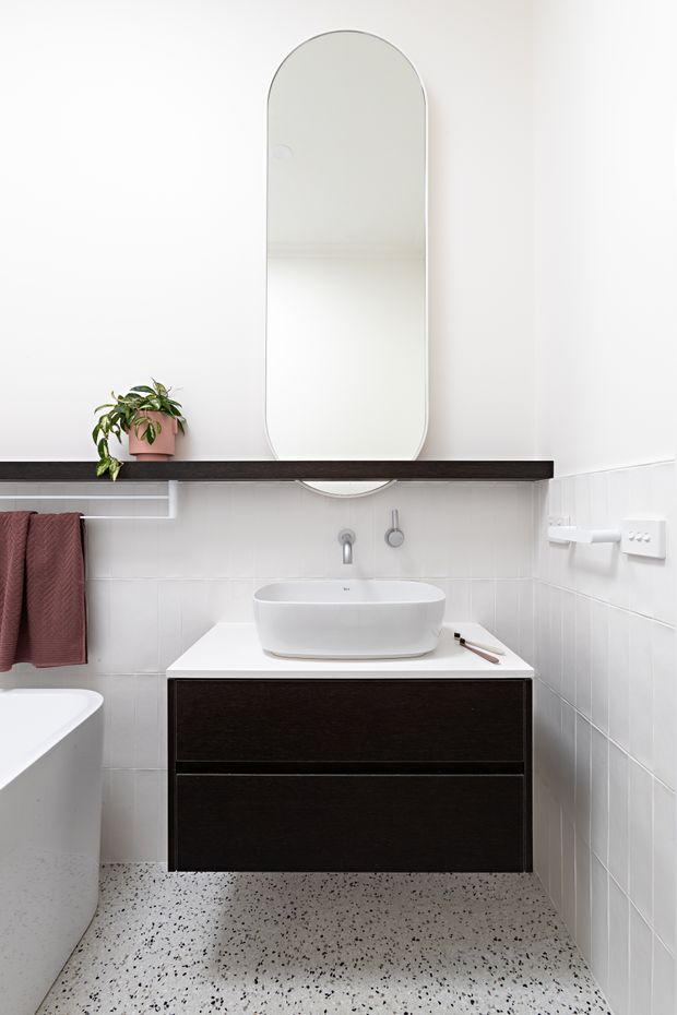A bright, compact contemporary bathroom featuring a white vessel basin on a floating dark timber vanity, a tall rounded-arch mirror, white tiled walls, and a terrazzo-style speckled floor. A freestanding white bathtub is partially visible at left, with a dark shelf, potted plant, and folded towel adding contrast.