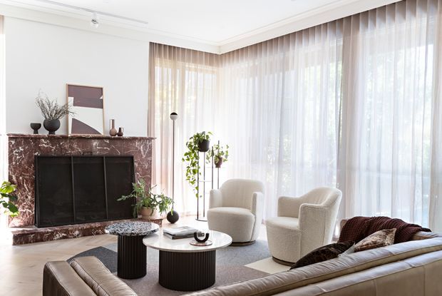 Bright, refined living room with a dramatic burgundy marble fireplace surround, white walls and ornate ceiling trim, full-height sheer curtains wrapping around corner glazing, and a contemporary seating arrangement. The room includes cream boucle lounge chairs, a dark leather sofa, sculptural round coffee tables, indoor plants, and restrained abstract artwork. The overall composition blends classic architectural detailing with contemporary furnishings.
