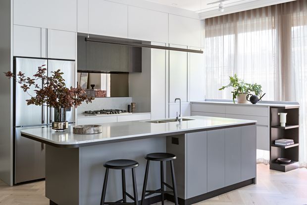 A bright, contemporary open-plan kitchen with a large central island, integrated white cabinetry, pale grey island panels, a stainless-steel refrigerator, gas cooktop, undermount sink, and slim linear pendant lighting. Two black stools provide island seating, while sheer curtains and indoor plants add softness to the clean, refined space.