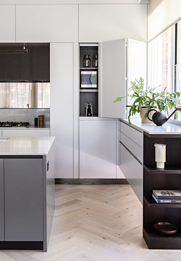 A bright, refined contemporary kitchen with full-height white cabinetry, a dark timber-toned cooking zone and open shelving, a pale stone island, integrated appliances, and a light herringbone timber floor. Large windows bring in abundant natural light, while black accents and potted greenery add contrast.