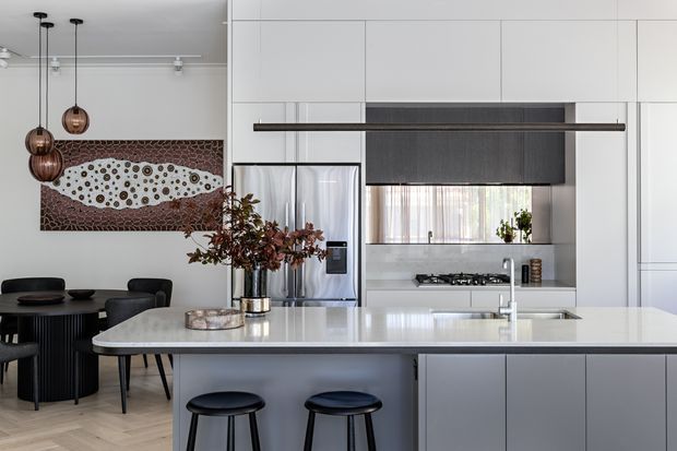 A refined open-plan kitchen and dining area with a large pale stone island, integrated white cabinetry, stainless-steel appliances, a dark timber-toned rangehood panel, and a gas cooktop beneath a window. Two black stools sit at the island, while a round dining table and chairs occupy the adjacent area. Pendant lights, contemporary artwork, and a floral arrangement add visual interest.