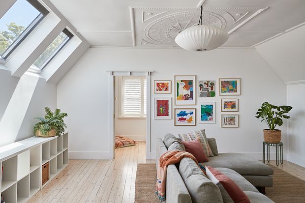 Bright attic-style living room with sloping roof glazing, white walls, pale timber floorboards, an ornate decorative pressed ceiling, and a large grey sectional sofa. A colourful gallery wall, indoor plants, woven textures, and a sculptural pendant add character. A doorway opens to a small adjoining bedroom or sleeping area.