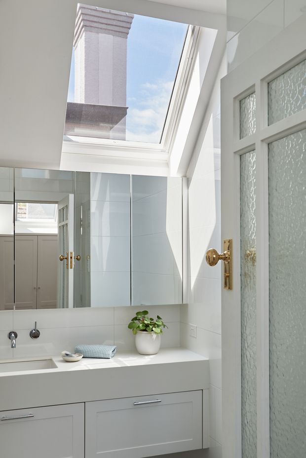 Bright, compact bathroom vanity area with white built-in cabinetry, a white countertop and integrated basin, large mirrored wall panels, white tiled walls, and a frosted textured-glass door with brass hardware. A large skylight brings in strong natural light and is bordered by white architectural trim. Reflections reveal additional cabinetry and the vanity area.