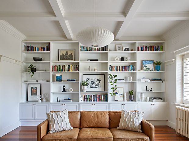 A bright, refined living room dominated by a full wall of custom white built-in bookcases and lower cabinetry filled with books, framed artwork, ceramics, and plants. A tan leather sofa with textured cushions sits in the foreground, beneath a large sculptural pendant light. The room features a white coffered ceiling, white panelled walls and trim, dark timber flooring, and a radiator beneath white plantation-style shutters.
