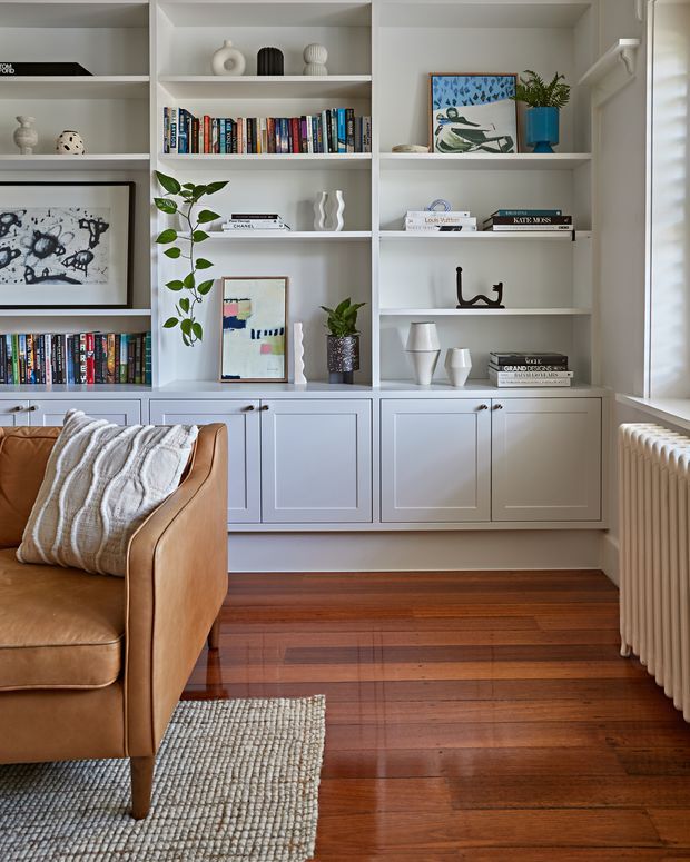 A bright, refined living room corner featuring a full wall of white built-in shelving and lower cabinetry, styled with books, ceramics, framed artwork, and indoor plants. A caramel leather armchair with a textured throw sits on a woven rug over polished dark timber flooring. A traditional white radiator is visible beside the window, adding character to the space.