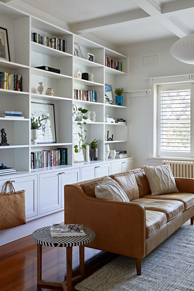 A bright, refined living room with extensive white built-in bookcases and lower cabinetry, filled with books and curated decor. A cognac leather sofa with textured neutral cushions faces a small round patterned coffee table. The room includes warm timber flooring, a woven area rug, white decorative ceiling beams and trim, and a large window with white horizontal louvres/shutters. Natural daylight creates a welcoming, balanced atmosphere.