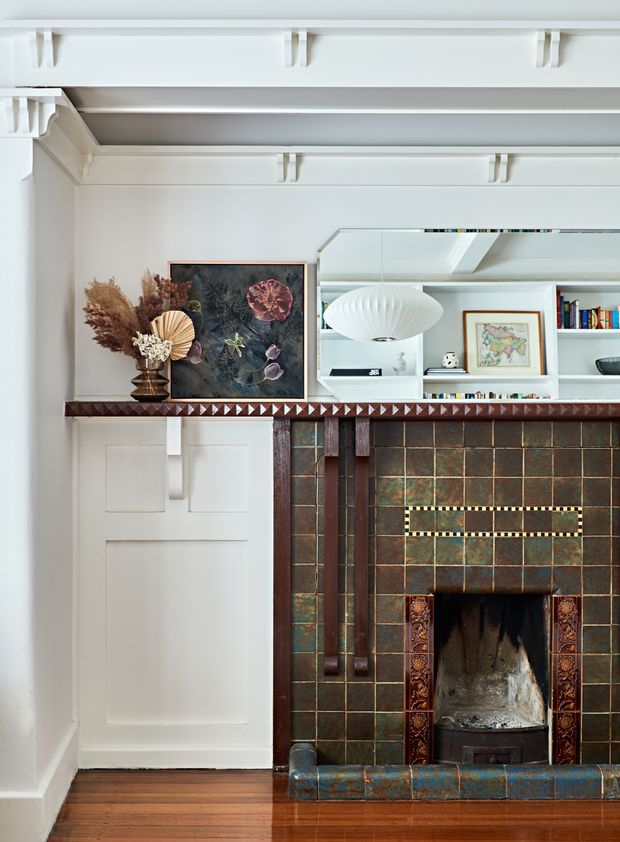 Interior detail of a refined traditional living room centred on an ornate tiled fireplace. The fireplace has dark, aged square tiles with decorative patterned surrounds, a carved dark timber mantel, and a raised tiled hearth. White built-in cabinetry and panelled wall detailing sit beside and above the fireplace, with open shelving, artwork, books, and a sculptural pendant reflected in a large mirror. Timber flooring and a dried floral arrangement add warmth.