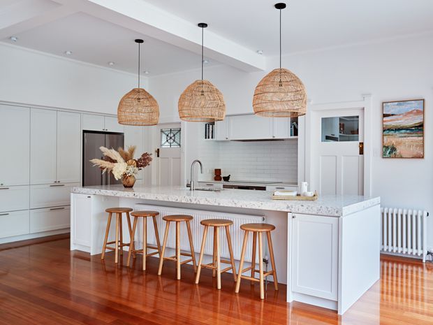 Bright, spacious white kitchen with a large island and six timber bar stools. The room features white shaker-style cabinetry, a pale stone benchtop, white subway-tile splashback, polished timber flooring, integrated appliances, a farmhouse-style sink, and three woven pendant lights. A radiator, framed artwork, and a glazed internal door are visible at the side.