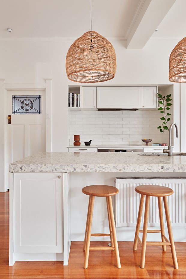 Bright, refined kitchen interior viewed from the living side of a central island. The space features white shaker-style cabinetry, a white subway-tile splashback, a pale terrazzo-like stone island benchtop, warm timber flooring, woven pendant lights, timber bar stools, and a stainless-steel tap. A decorative glazed entry door and indoor plant are visible in the background.