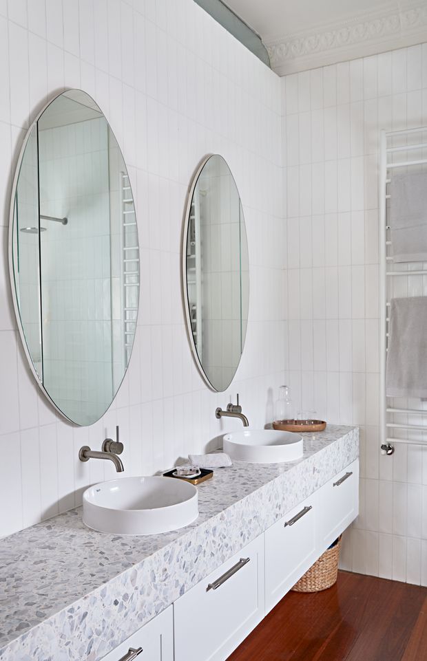 Bright, refined bathroom with a double vanity, white vessel basins, wall-mounted brushed metal taps, a pale terrazzo-style benchtop, white tiled and vertically panelled walls, oval mirrors, ornate cornice detailing, white cabinetry, and a heated towel rail. Dark timber flooring adds warmth and contrast.