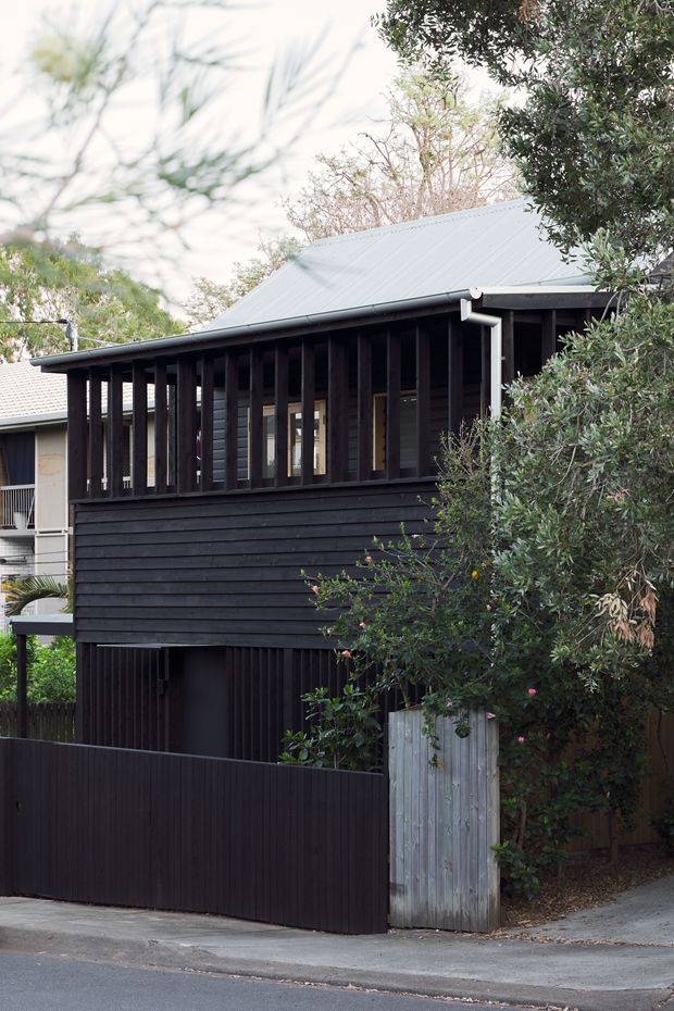 Street-facing view of a compact two-storey dark-clad house with a steep pitched metal roof, horizontal timber wall boards, and a partially enclosed upper-level verandah or balcony screened by vertical timber battens. A dark pedestrian gate and fence define the frontage, with mature vegetation surrounding the building.