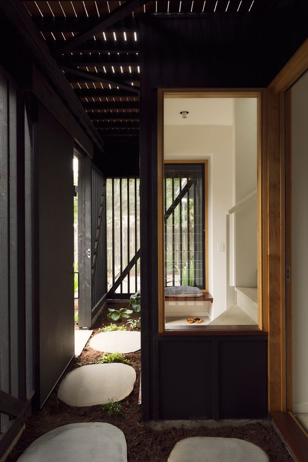 A narrow, dark timber-lined entry passage with large, pale stepping stones set through a planted ground surface. Black timber framing and slatted overhead elements create a screened, semi-outdoor corridor, while a glazed opening reveals a bright interior stair landing and a timber-framed doorway. The composition contrasts deep charcoal and brown timber with warm white interior surfaces and natural greenery.