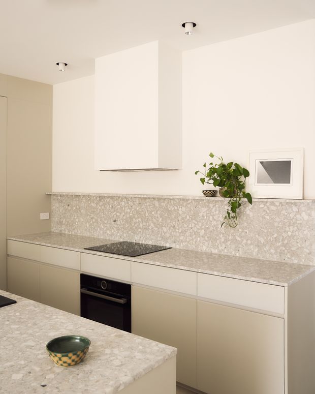 A refined contemporary kitchen with a pale terrazzo stone benchtop and matching full-height splashback. Soft beige flat-panel cabinetry runs along the rear wall, with an integrated black cooktop and oven beneath a minimalist white rangehood. A foreground island repeats the terrazzo finish, while recessed ceiling spotlights, a potted trailing plant, and a framed artwork complete the calm, neutral space.
