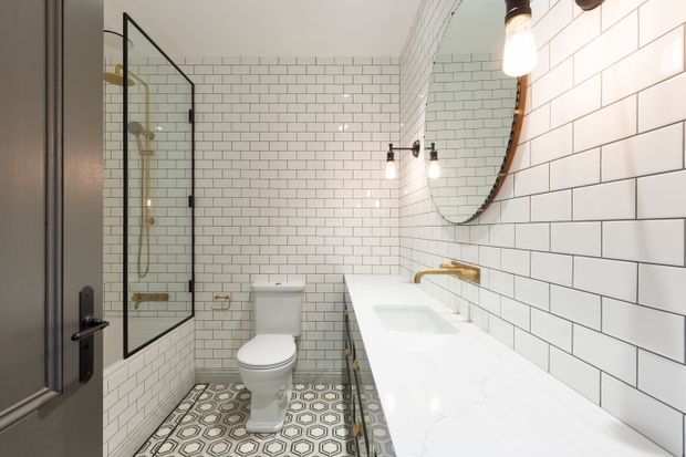 Bright, narrow bathroom with white subway-tiled walls, a long white vanity with integrated basins and brass fittings, a black-framed glass shower enclosure, wall-mounted exposed-bulb lighting, and patterned geometric floor tiles. A toilet and large round mirror complete the compact layout.